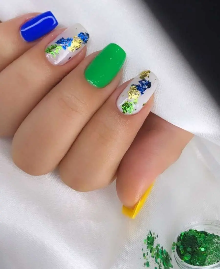 Imagem de mão decoradas com esmalte.  Cores usadas, verde, amarelo, azul e branco. Polegar: amarelo. Indicador: branco com glitter verde, dourado e azul. Dedo Médio, verde. Anelar: branco com glitter verde, dourado e azul. Mínimo: azul.