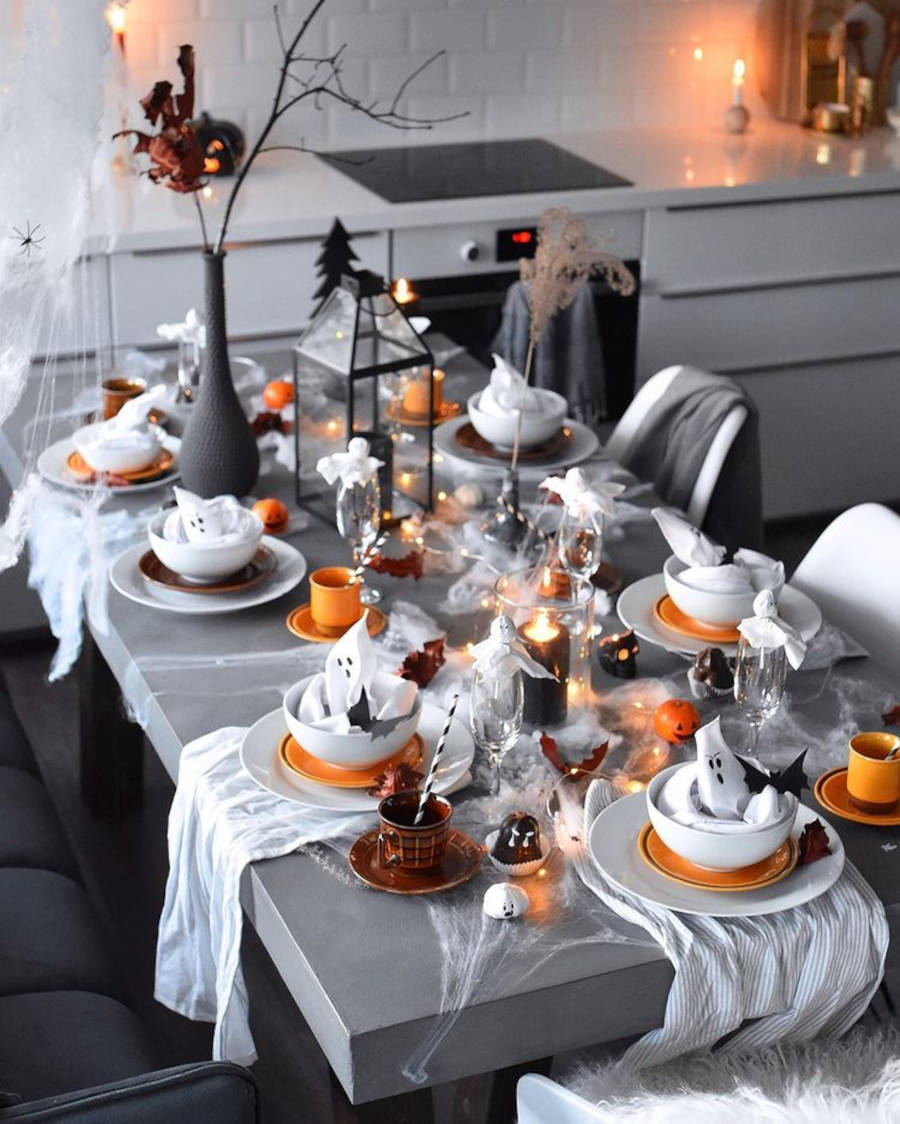 Mesa posta de halloween com teias falsas, velas de caveira, fantasmas de papel no bowl, xícaras laranjas e mais