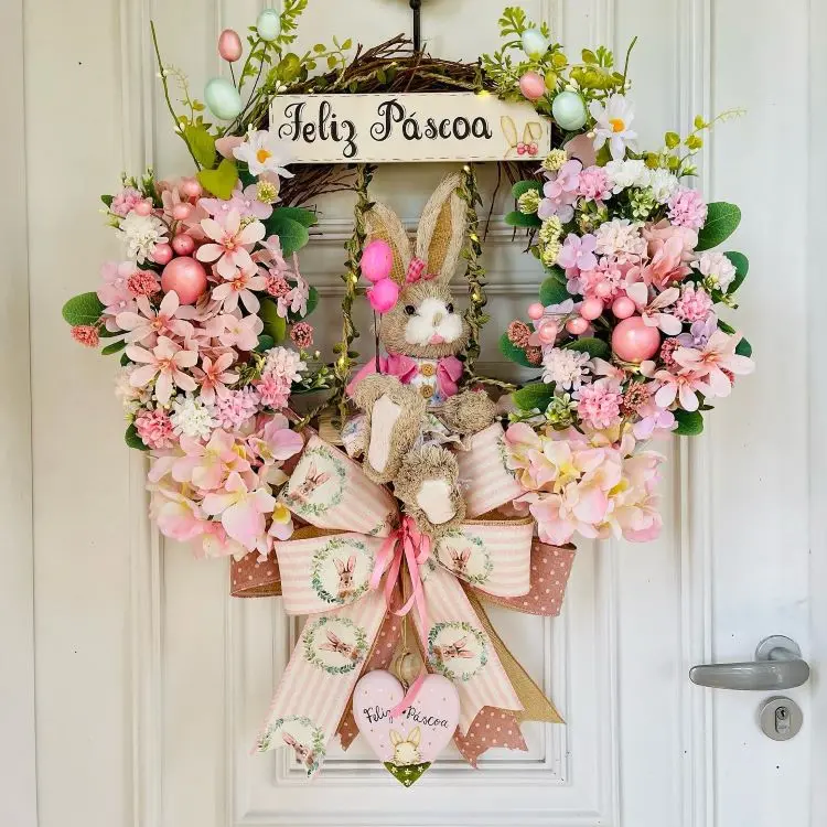 Guirlanda de Páscoa decorativa com flores rosa, coelhinho central, ovos coloridos e laço com detalhes estampados. Placa superior com “Feliz Páscoa” e enfeites pendurados. Fundo branco, design delicado e acolhedor, ideal para celebração pascal.