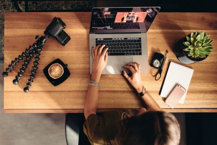 Vista aérea de uma pessoa editando um vídeo em um laptop, cercada por equipamentos como câmera e tripé, simbolizando o trabalho na faculdade de influencers. Há uma xícara de café, um caderno e um celular sobre a mesa de madeira.