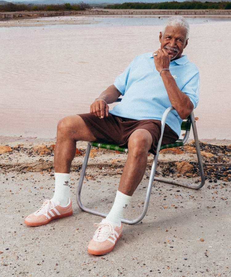 Foto de homem sentado em cadeira de praia usando camiseta azul, short marrom, meia branca e tênis da parceria de Bad Bunny com adidas