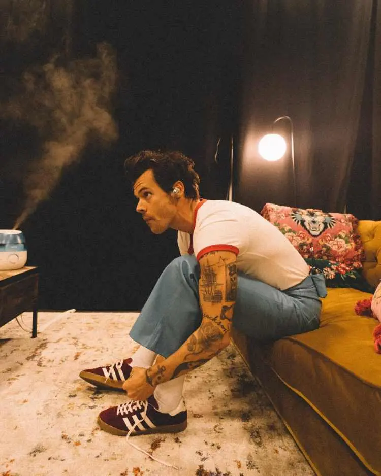 Harry Styles sentado em sofá amarelo, está vestindo camiseta branca com golas vermelha e calça jeans, com braço tatuado à mostra