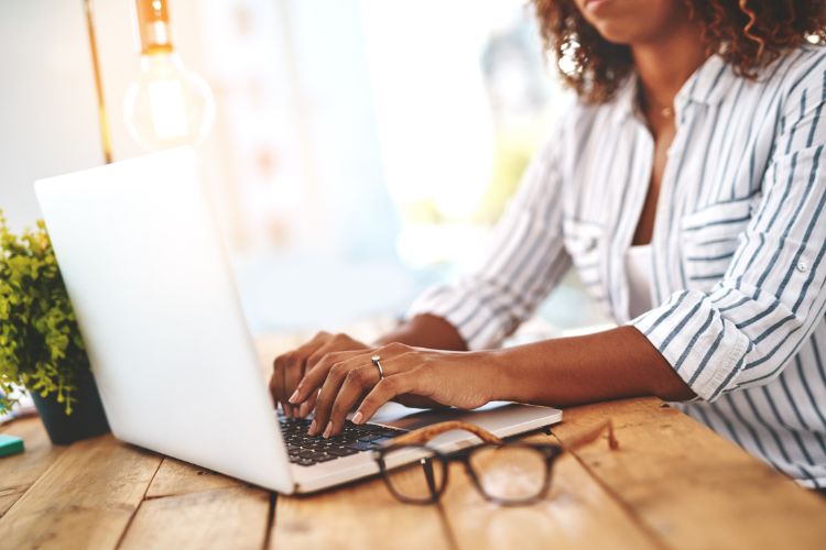 Mulher trabalhando em um laptop em uma mesa de madeira, ao lado de um vaso de planta e óculos. O ambiente iluminado e descontraído reflete produtividade, foco e equilíbrio entre trabalho e conforto em um espaço inspirador e moderno.





