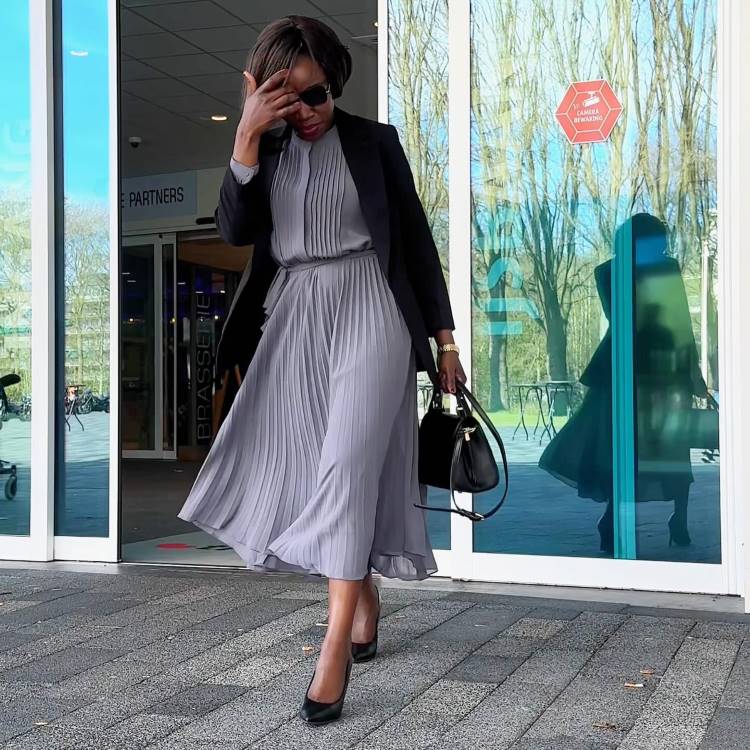 Mulher elegante caminha em frente a um prédio com fachada de vidro. Usa vestido plissado cinza, blazer preto e sapatos de salto alto pretos. Segura bolsa preta estruturada e usa óculos de sol. Perfeita referência para looks de trabalho sofisticados.





