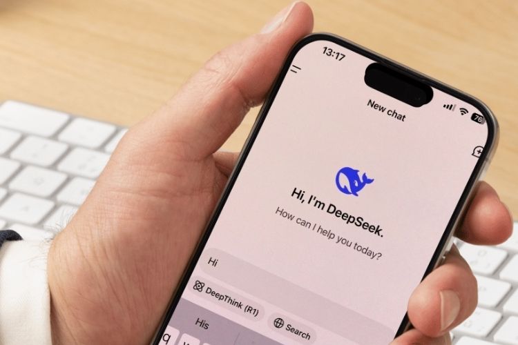Uma mão segura um smartphone exibindo a interface do chatbot "DeepSeek" com um logotipo azul de baleia. Ao fundo, há um teclado branco e uma superfície de madeira clara que compõem o ambiente.