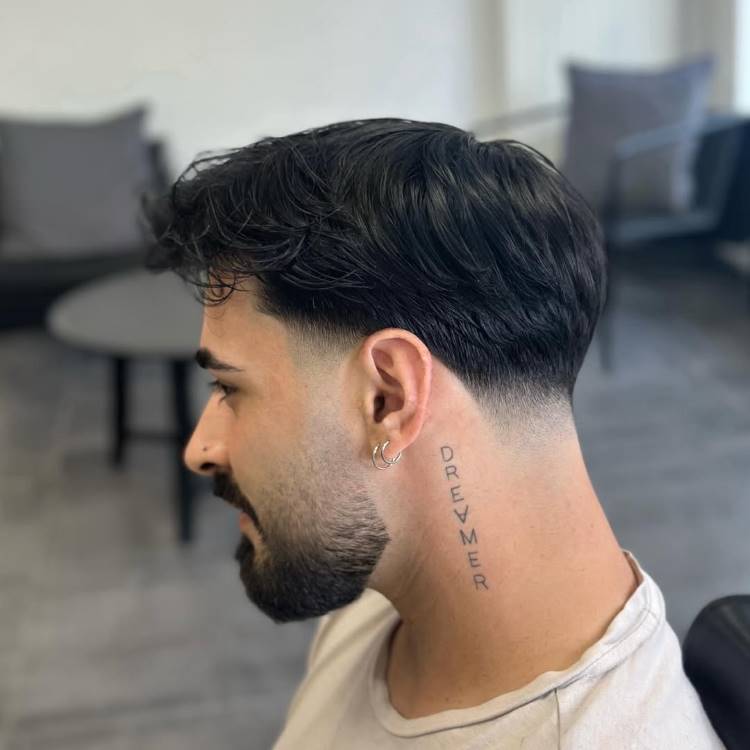 Um homem com barba, de perfil, e um corte de cabelo mid fade. O cabelo do topo é mais longo e escuro, com textura ondulada. As laterais e a nuca têm um degradê que se conecta à barba.