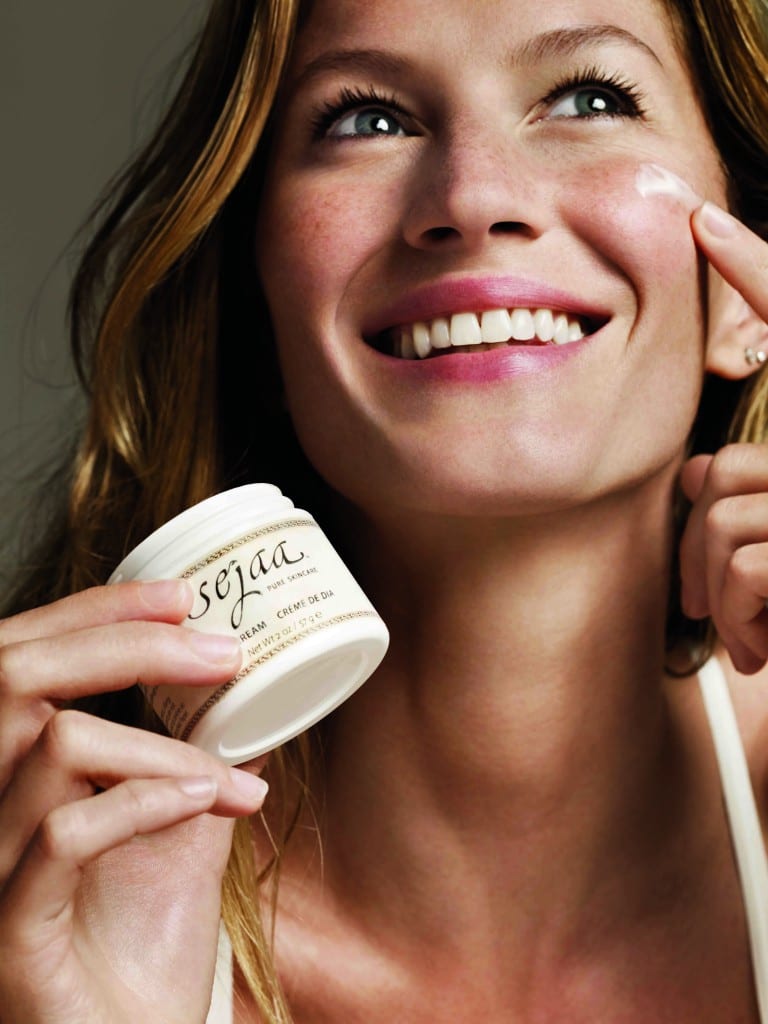 Gisele Bündchen sorrindo e passando creme de sua marca