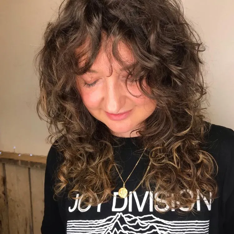 uma mulher branca de cabelos enrolados com uma camiseta da banda joy division, usando um corte de cabelo wolf moreno iluminado