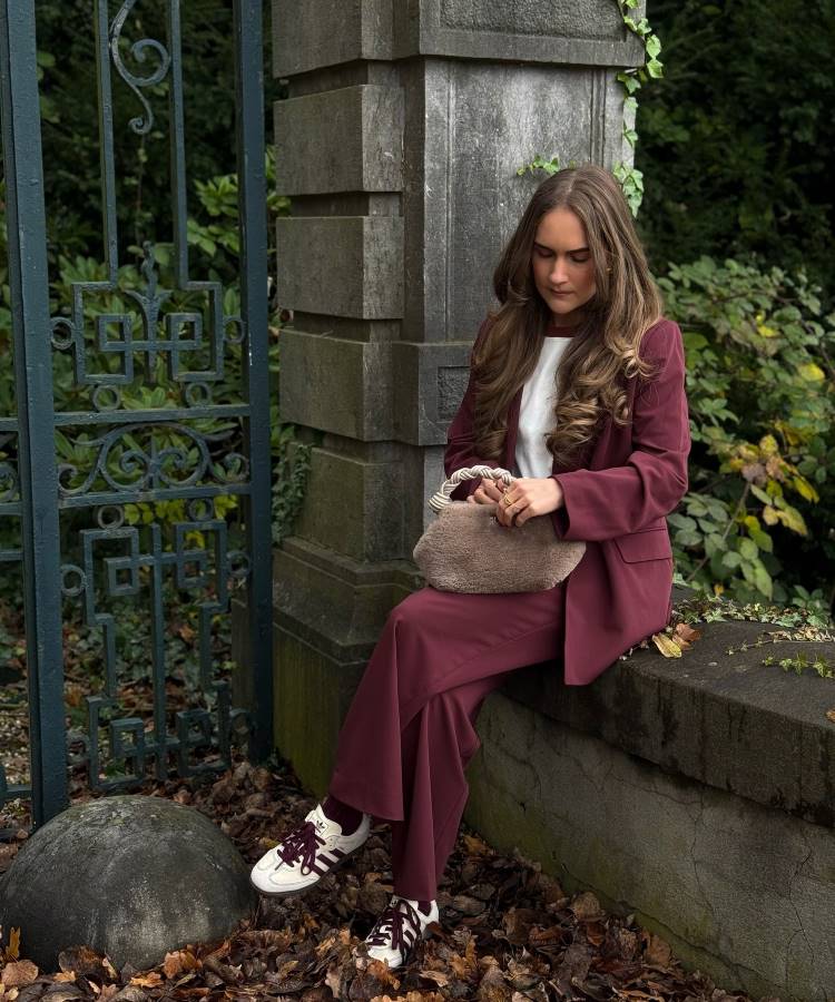 Mulher sentada em ambiente externo com blazer e calça vinho, camiseta branca e tênis esportivo combinando. Segura bolsa de mão peluda bege. Vegetação e muro clássico ao fundo. Inspiração casual e moderna para looks de trabalho com conforto e estilo.





