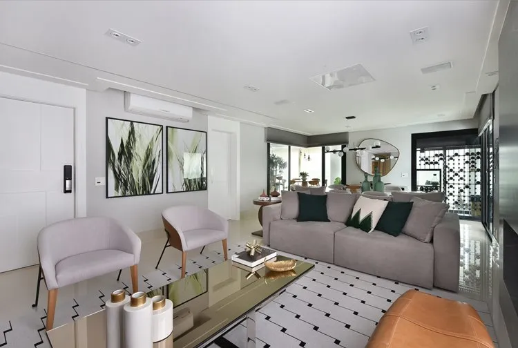O sofá e poltronas são cinza, as almofadas são cinza e verde escuro, o quadro apresenta uma planta verde e o tapete é cinza e preto.