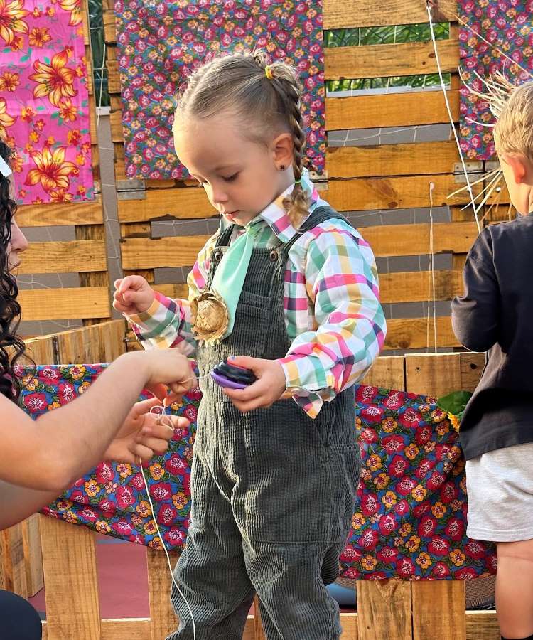 Menina usando camisa xadrez com jardineira verde em festa junina.

