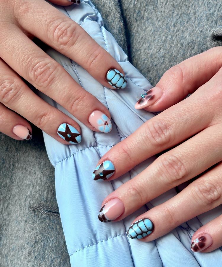 Mãos destacando estilos de unhas curtas, decoradas com esmalte nude e detalhes em azul e marrom, incluindo flores, estrelas e padrões geométricos. O design criativo combina tons suaves com contraste marcante. Fundo com tecido azul acolchoado, criando um visual harmonioso.