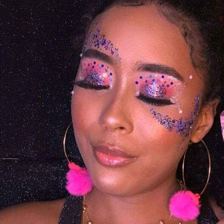 Mulher de pele negra com maquiagem rosa e glitter roxo de diversos tamanhos nos olhos, acima da sobrancelha e abaixo dos olhos 