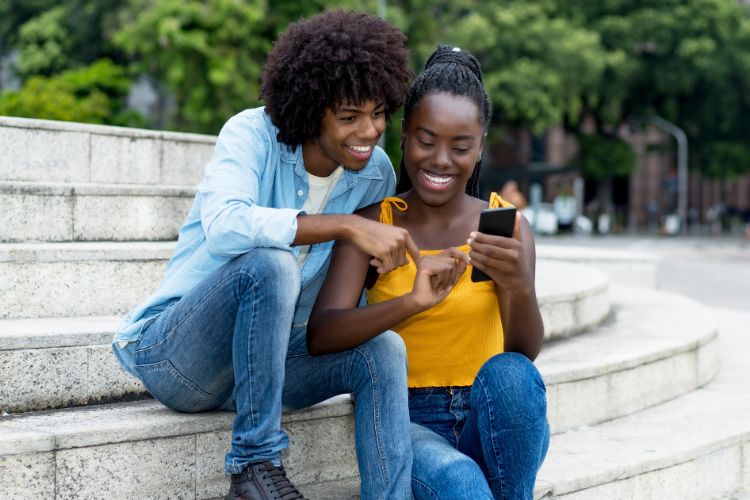Dois jovens sentados em escadaria ao ar livre, sorrindo enquanto olham para um celular. O rapaz aponta para a tela, sugerindo estarem discutindo algo, possivelmente sobre redes sociais ou explorando o tema “Quanto custa um publi?” em influenciadores.