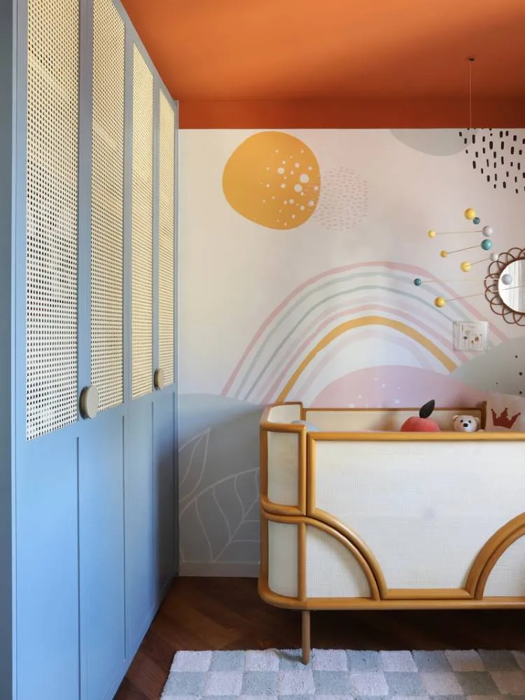 Esta imagem mostra um quarto de bebê com um teto colorido laranja vibrante. Há um berço de madeira clara com tela de vime e um mural de parede com arco-íris e formas abstratas. Um guarda-roupa azul com portas de vime adiciona um toque de charme único ao ambiente.

