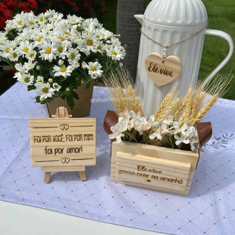 Arranjo com enfeites de Páscoa, incluindo placa de madeira com frase religiosa, jarro branco com coração de madeira gravado “Ele vive” e mini caixote decorado com flores e trigo. Toalha de mesa branca e margaridas ao fundo.