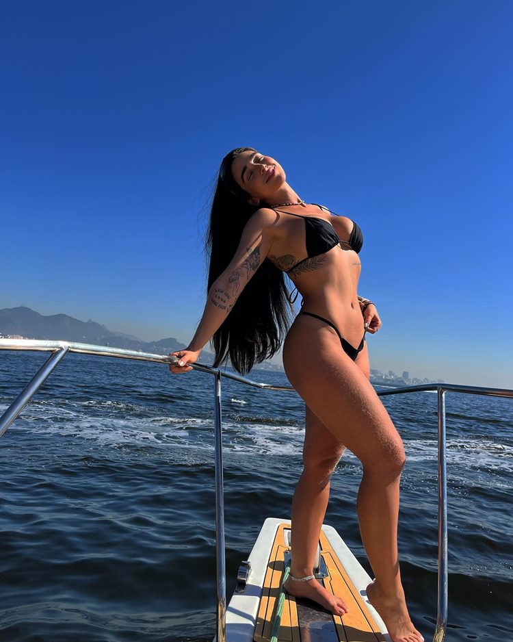Participante do BBB 24 Giovanna Lima de biquíni preto em embarcação em alto mar, com montanhas no fundo e céu azul