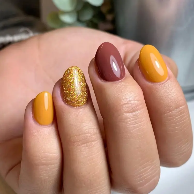 Mão de pele clara com unhas arredondas curtas em mostarda, marrom e dourado 