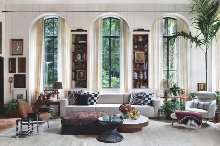 Sala com sofá cinza de almofadas brancas e pretas, poltronas, grandes janelas, quadros, livros e mesas de centro 