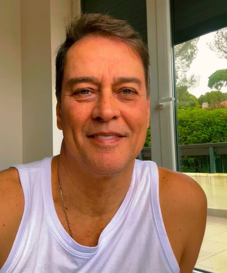 Um homem de meia-idade, com cabelos castanhos claros e olhos verdes, sorri levemente para a câmera. Ele usa uma regata branca e uma corrente no pescoço. O fundo mostra uma janela com plantas do lado de fora.
