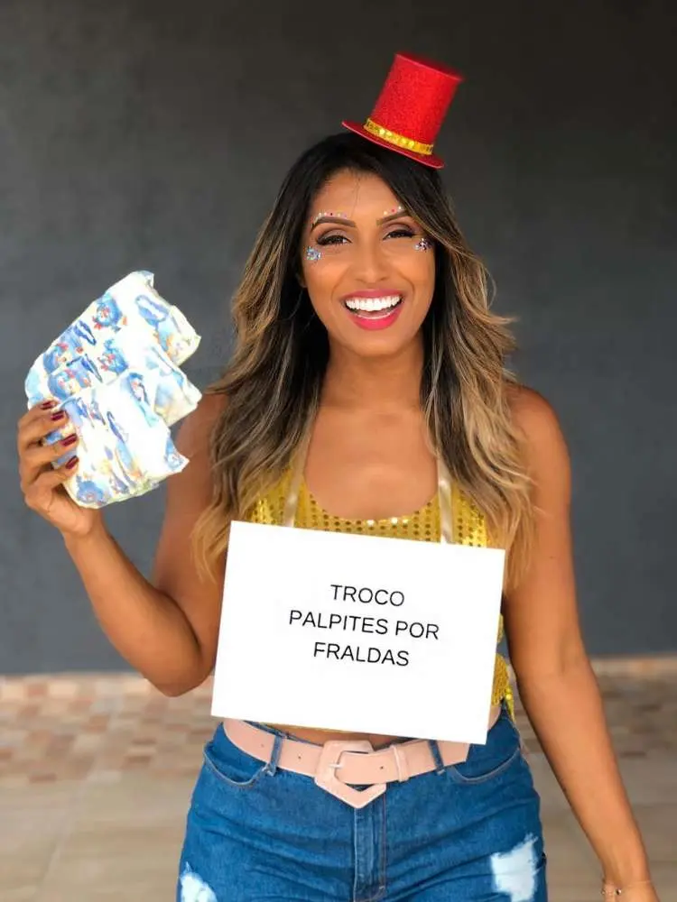 Foto de mulher de pele morena usando tiara de cartola vermelha, top dourado, shorts jeans e plaquinha escrito "troco palpites por fraldas"
