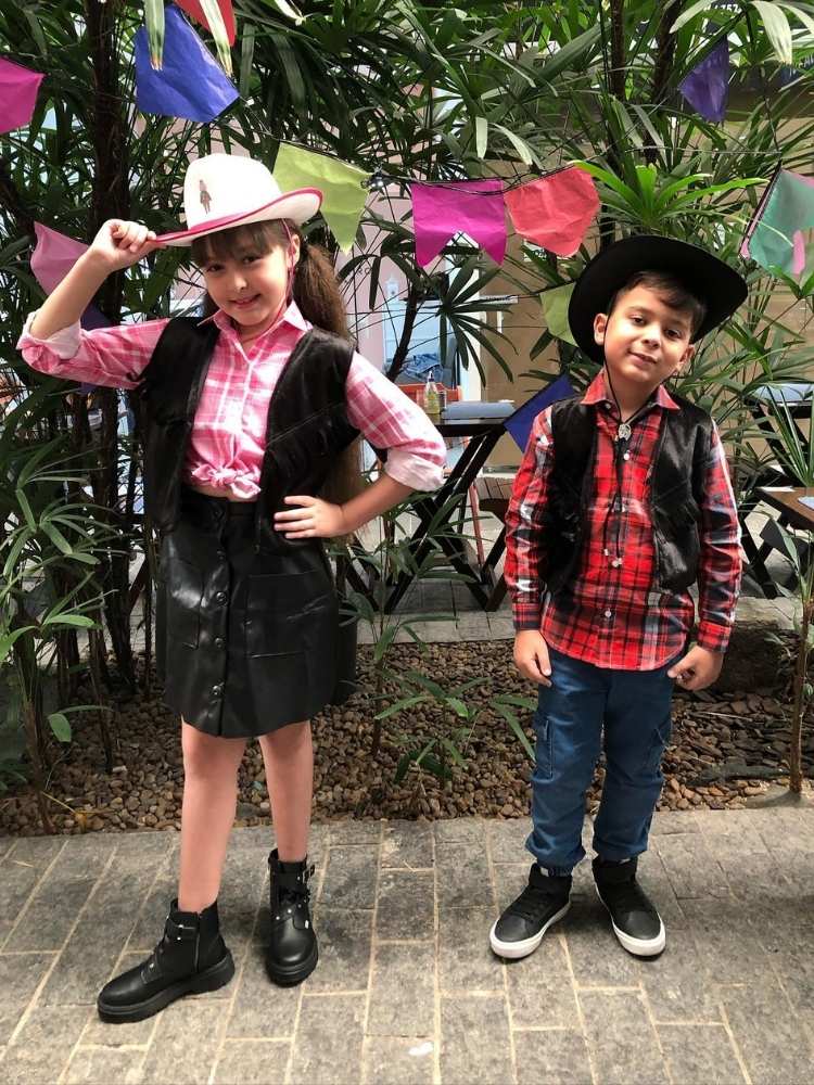 Menina usando camisa xadrez rosa, colete preto, saia preta, bota e chapéu country. Menino usando camisa xadrez vermelha, colete preto, chapéu e country preto