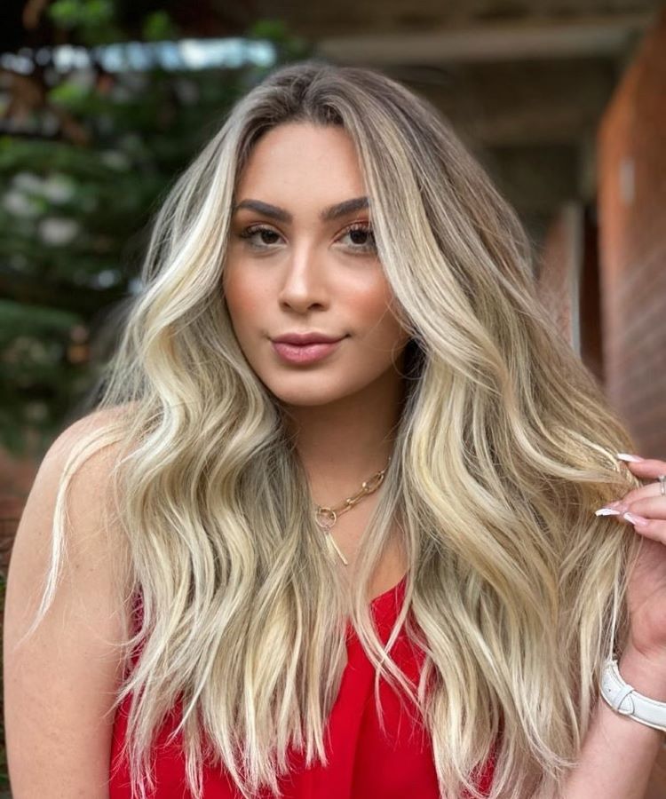 Mulher com cabelos longos e ondulados, exibindo uma das tendências atuais de cores de cabelo: loiro iluminado com mechas em tons dourados e platinados. Ela usa maquiagem leve, veste uma blusa vermelha e está em ambiente externo arborizado.