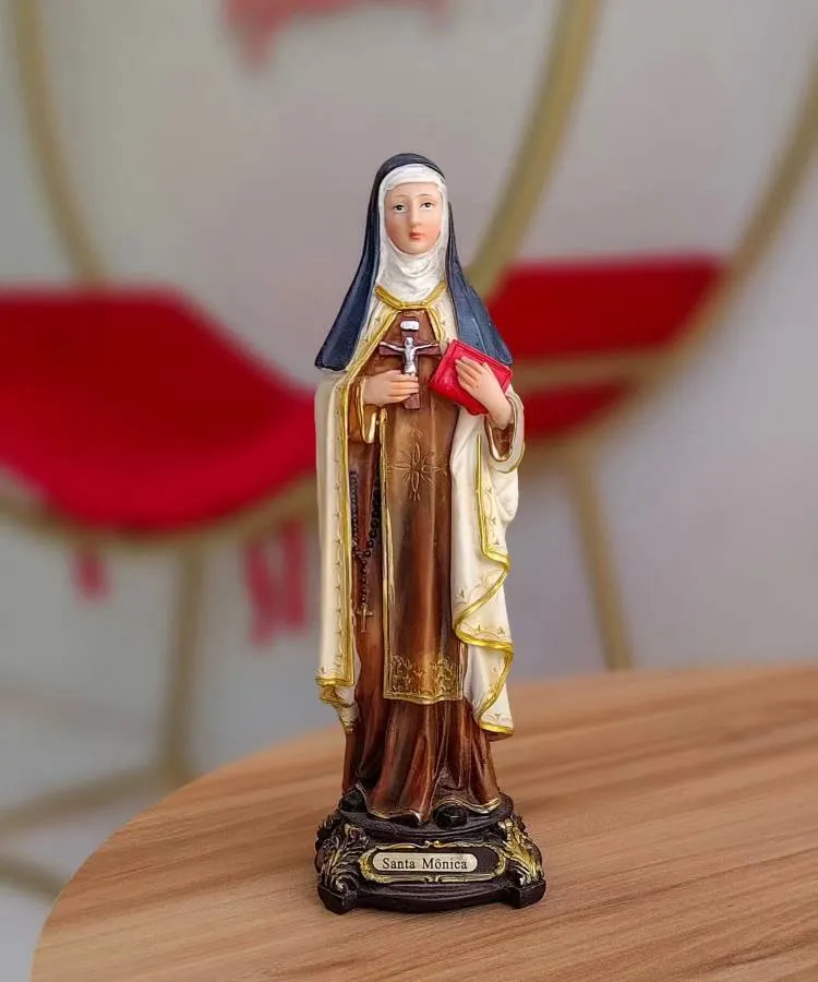 A imagem mostra uma estátua de Santa Mônica. Ela está vestida com um hábito religioso, composto por túnica marrom, manto branco e véu preto. Santa Mônica segura um livro vermelho e um rosário. A base da estátua tem uma placa com o nome "Santa Mônica". Ao fundo, há um ambiente com tons de vermelho e dourado.


