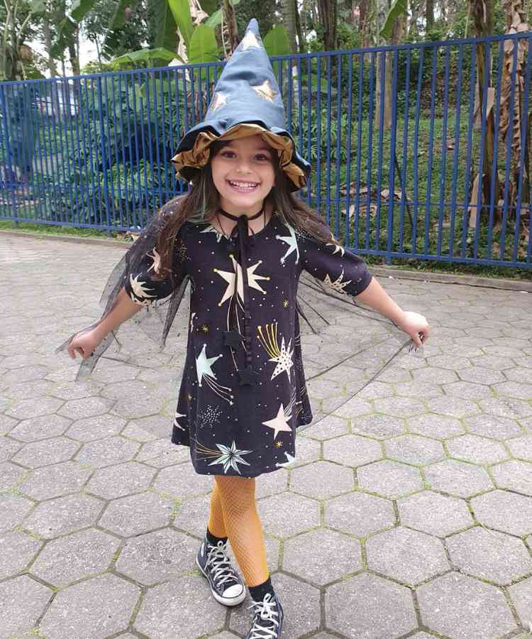 Garota com fantasia de bruxa infantil. Cores predominantes: preto, branco e dourado. Usa: vestido, capa de tule, chapéu pontudo, meia calça arrastão, tênis