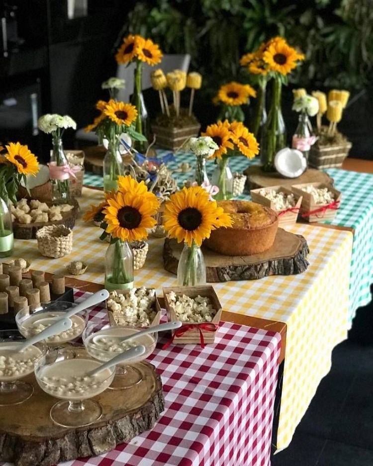 A imagem representa decoração para festas juninas com flores. Contém mesa com toalhas xadrez vermelha, amarela e verda, com comidas como pipoca e canjica