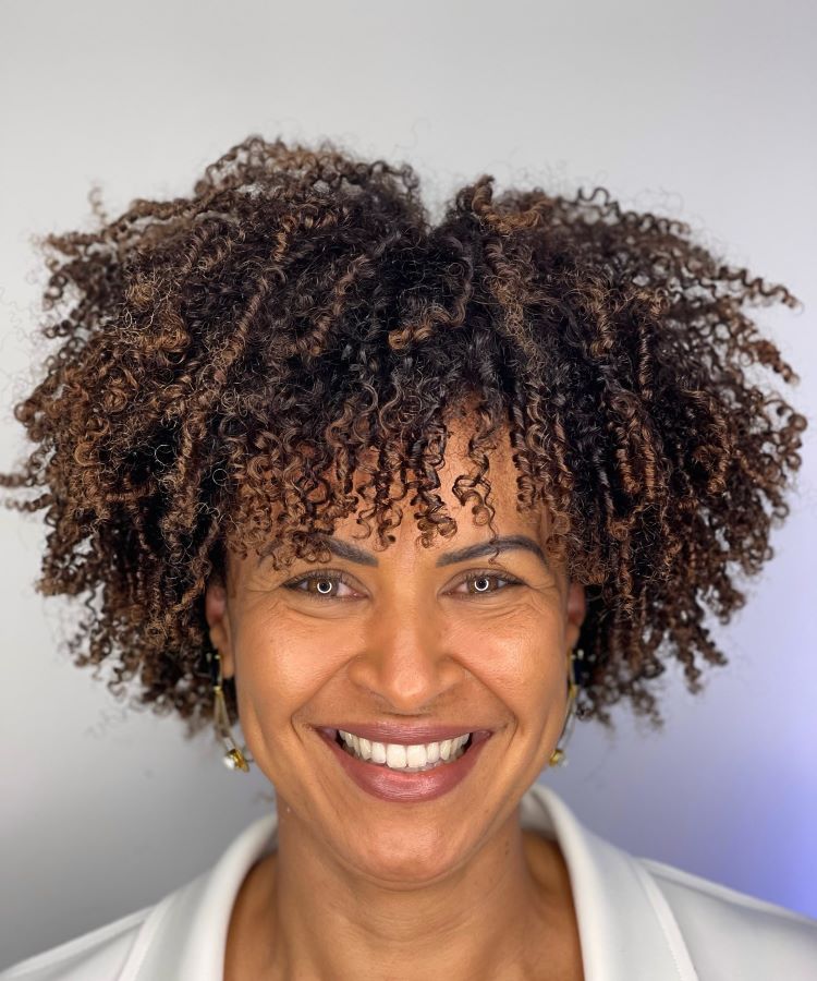 Mulher com cabelo curto e cacheado, destacando cores de cabelo modernas com tons castanhos e mechas em nuance caramelo. Ela sorri, veste blazer branco, usa brincos delicados e está em um ambiente iluminado com fundo neutro e suave.