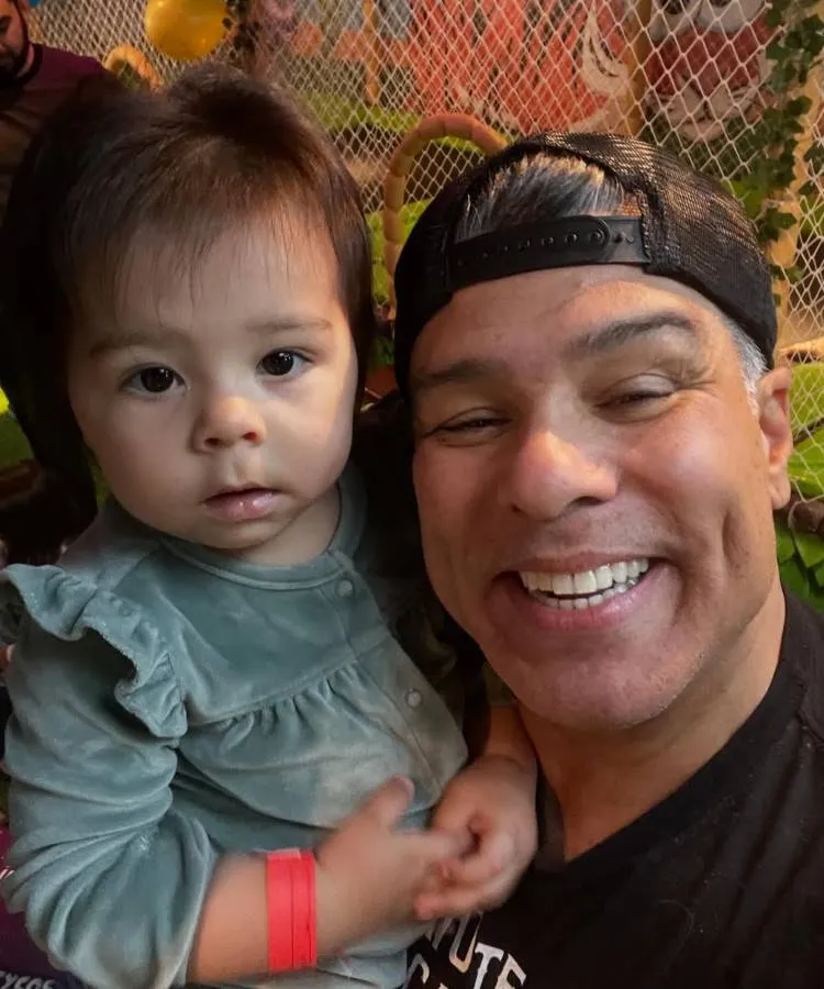 Um homem de boné preto e sorriso largo segura um bebê com cabelo escuro e roupinha verde-acinzentada, que olha para a câmera. Nomes diferentes de filhos de famosos: a pulseira vermelha no pulso do bebê sugere um evento ou identificação, como em parques ou festas.

