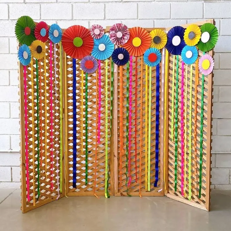 Um biombo de madeira decorado com fitas coloridas verticais e leques de papel em tons vibrantes no topo. Um cenário festivo para uma celebração ou evento.