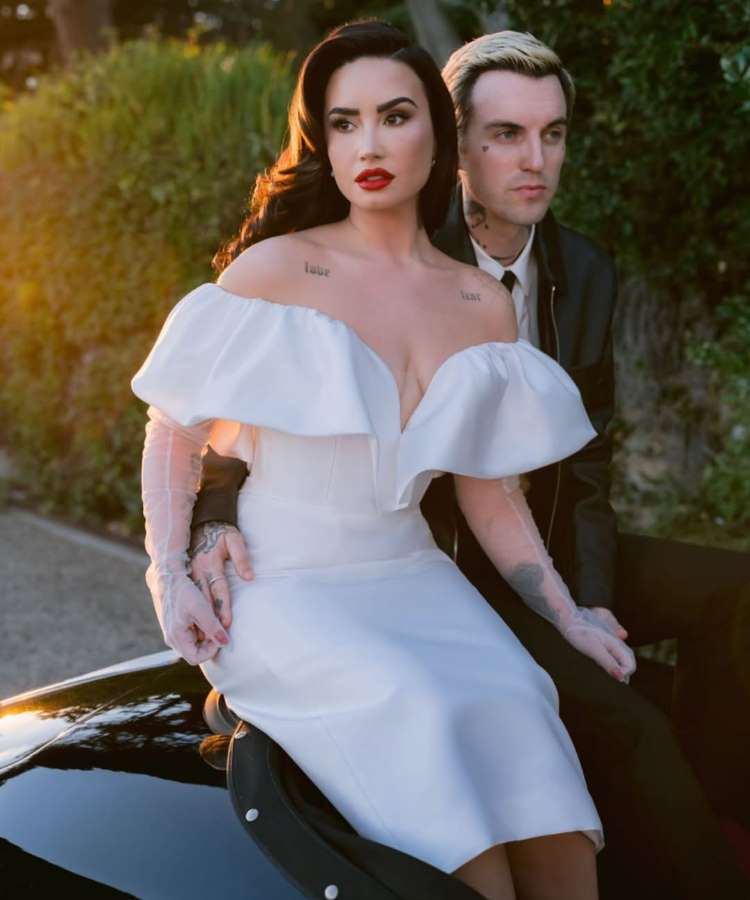 Demi Lovato, de vestido branco de ombros caídos e luvas transparentes, senta-se no capô de um carro, olhando para a câmera. Ao lado dela, um homem de cabelo loiro e jaqueta de couro está atrás dela. A luz dourada do sol poente ilumina a cena.