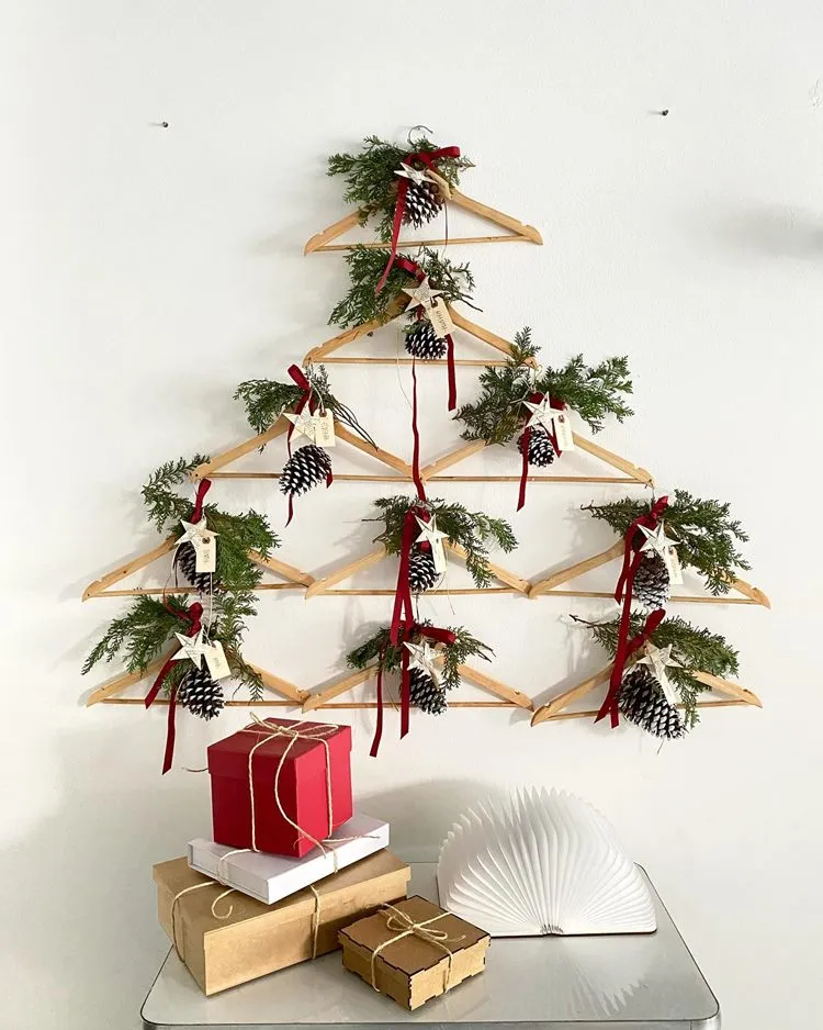 Árvore-de-Natal de parede feita com cabides e decorada com pinhos. Abaixo, mesa com caixas de presente