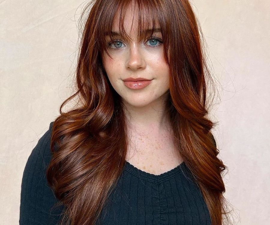 Mulher com cabelos longos ruivos monocromáticos, levemente ondulados e com franja suave. Ela usa maquiagem leve, com batom nude brilhante, e veste uma blusa preta texturizada. O fundo neutro em tom pastel realça os fios vibrantes e brilhantes.