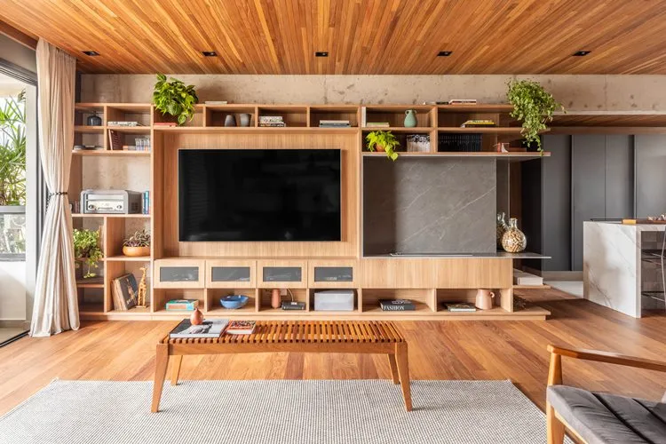 Vista ampla do home-theater do apartamento de 187 m² decorado. Móvel de madeira com várias repartições decorado com livros, caixas, plantas e outros objetos. Uma TV grande está ao lado da lareira