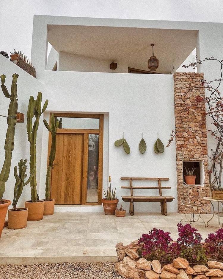 Imagem de fachada de casa simples branca decorada com pedras, cactos em vasos e pendurados na parede, banco e pequena mesa