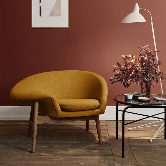 Sala com parede vinho e poltrona mostarda, além de mesa de vidro com planta fake e luminária