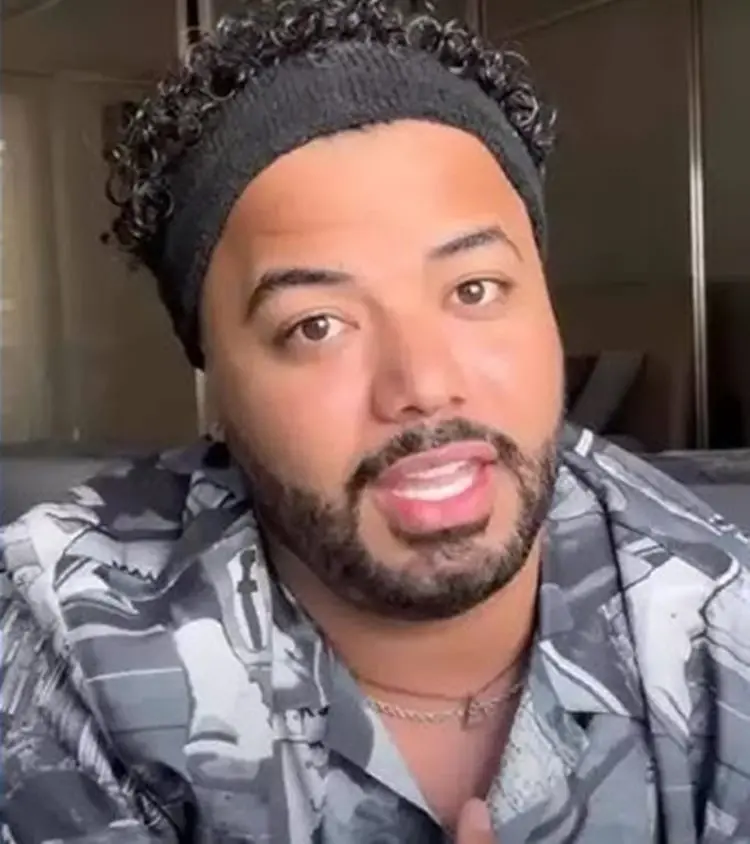 Um homem de barba e cabelo encaracolado, usando uma faixa de cabelo cinza e uma camisa estampada, olha diretamente para a câmera com a boca ligeiramente aberta. O fundo está desfocado.