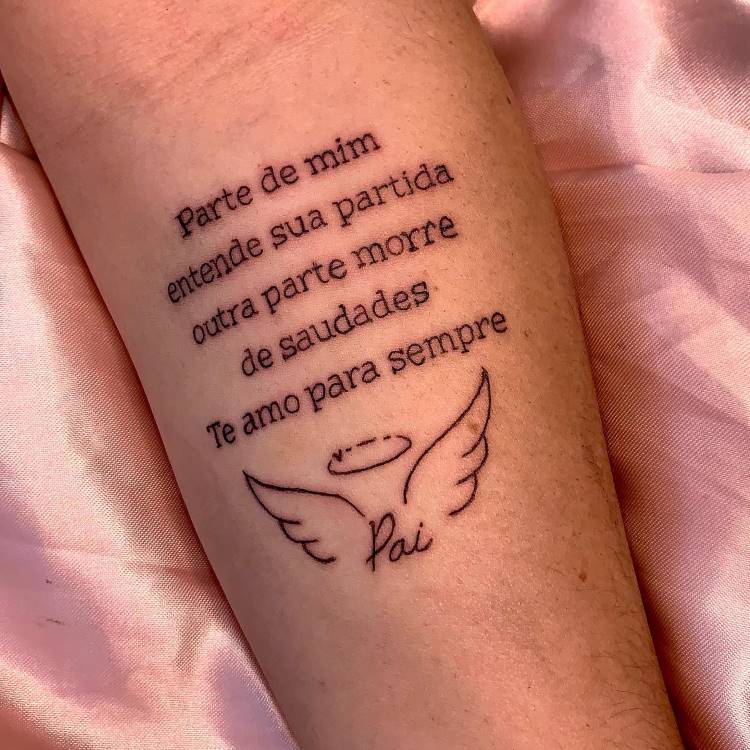 A imagem mostra uma tatuagem no antebraço com a frase: "Parte de mim entende sua partida outra parte morre de saudades Te amo para sempre". Abaixo, há asas de anjo com uma auréola e a palavra "Pai". A tatuagem é em preto e o fundo é um tecido rosa.







