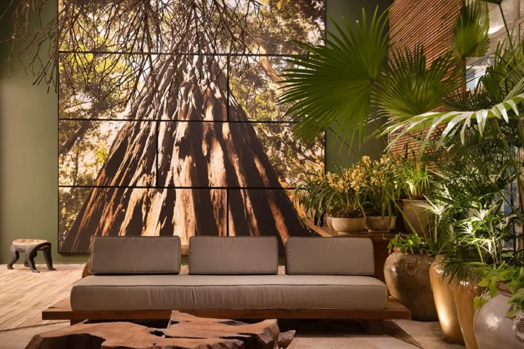 A imagem mostra uma sala de estar com um grande mural fotográfico de uma árvore imponente na parede traseira, mobiliário moderno e plantas vivas que complementam o tema natural.

