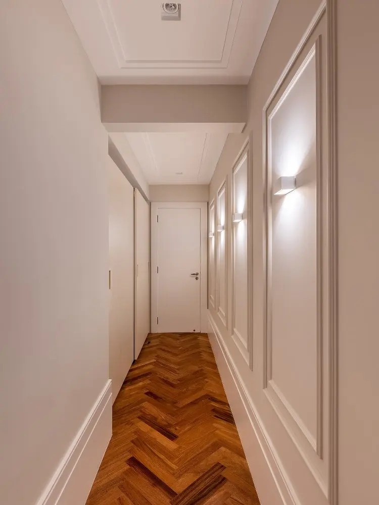 Corredor com piso de madeira, paredes brancas com  molduras e lustres dentro da moldura