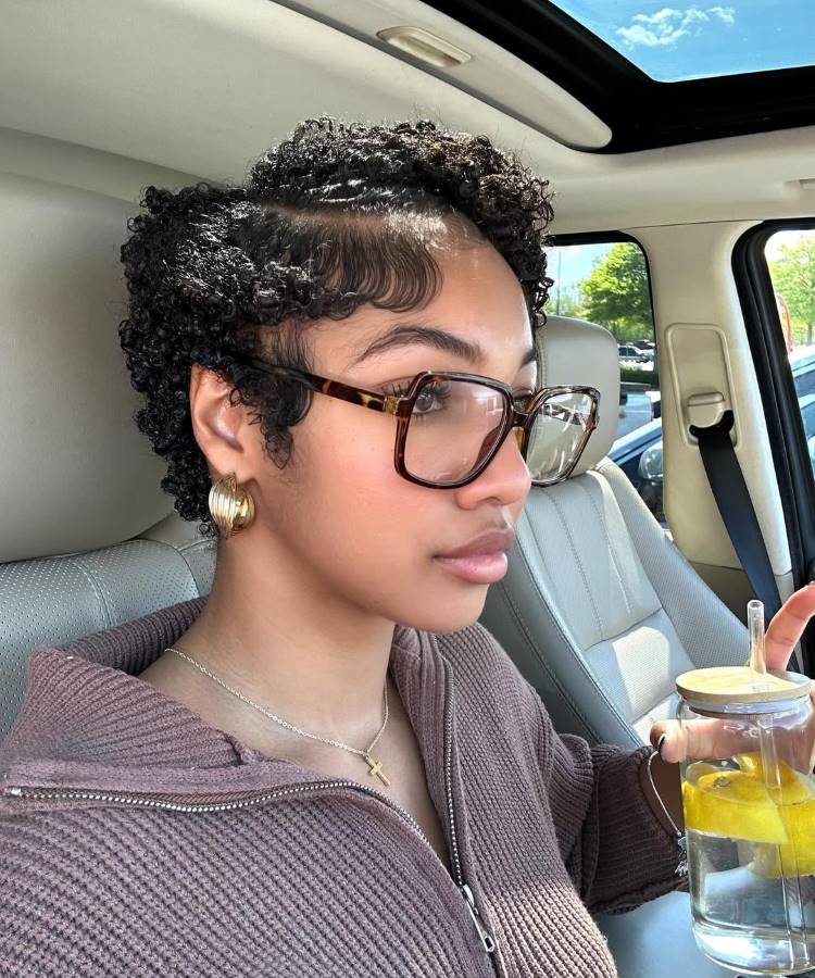 Uma mulher em um carro com um corte de cabelo cacheado curto e penteado para o lado. Ela usa óculos grandes, brincos de argola, e segura um copo com fatias de limão.