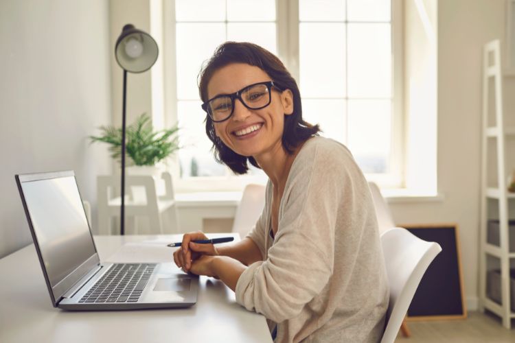 Mulher jovem, sorridente, de óculos, sentada em uma mesa branca com laptop, caneta e caderno. Ambiente iluminado por janela ampla, com planta decorativa ao fundo. Ela utiliza um verificador de plágio para garantir a autenticidade do conteúdo.