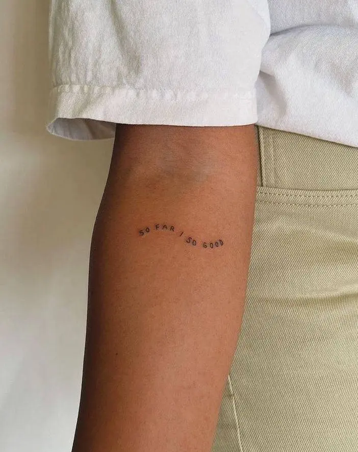 Tatuagem de frase delicada no braço escrito "so far/so good"