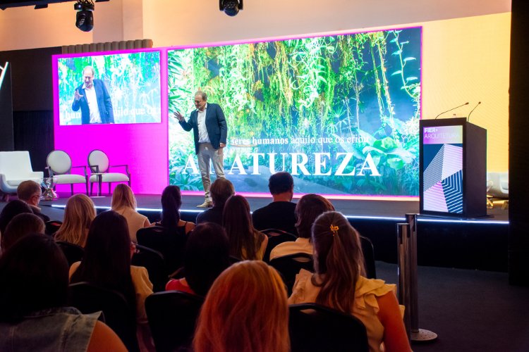 Alex Allard sobre palco preto palestrando para a plateia sentada e com foto de floresta e escrita "natureza" atrás do empresário