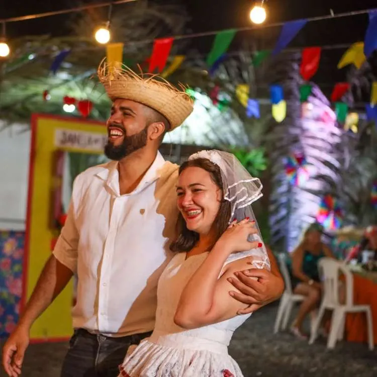 Um momento alegre capturado à noite mostra um casal se abraçando e rindo. O homem usa chapéu de palha e camisa branca, enquanto a mulher tem um véu e um vestido branco com detalhes em vermelho. Luzes e bandeiras coloridas enfeitam o fundo, indicando uma celebração animada ou evento pré-casamento.