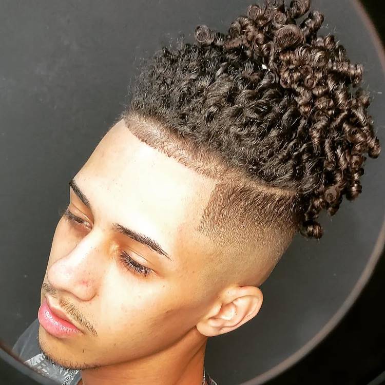 Um homem jovem, de perfil, com cabelo crespo e cacheado no topo da cabeça. Ele tem um high fade nas laterais e na nuca, com uma linha de design raspada para separar a parte de cima.







