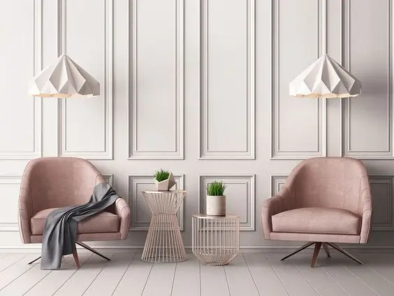 Sala com parede branca com as molduras, dois lustres brancos, duas poltronas rosas claras e dois apoios vazados com vaso 